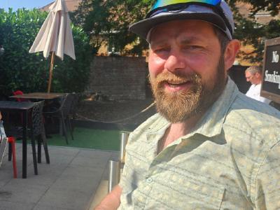 Steve Abraham with Peter Treviss at Battesford Court before the départ. Two cyclists resting at outdoor pub patio; left cyclist wears blue cap, sunglasses and black kit with red gloves; right cyclist has beard, cap with sunglasses on brim; tables and umbrellas behind them.
