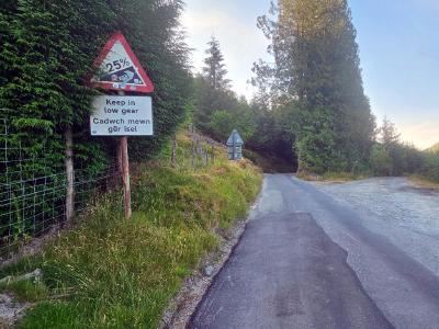 The Devils Staircase, Abergwesyn Road Steep hill warning sign showing 25 percent gradient with bilingual text Keep in low gear Gadwch mewn gear isel beside tree-lined country road curving into conifer forest.