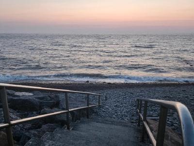 Aberaeron Pebble beach at dawn with metal handrails leading down to gentle waves, wooden groynes along shoreline, soft pink and orange sky gradient over calm sea.
