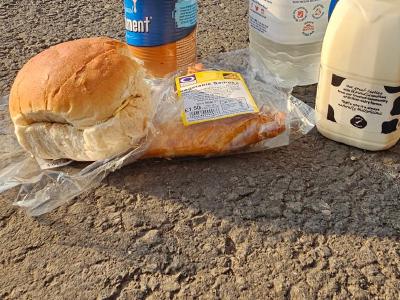 Breakfast near Llandeilo Cycling provisions on gravel ground including bread roll, baked potato, canned beans, large water bottle and milk carton, red hatchback car and houses in background.