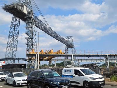 Newport Transporter Bridge Industrial transporter bridge with steel lattice towers and suspended gondola structure spanning overhead, cars parked in foreground under blue cloudy sky.