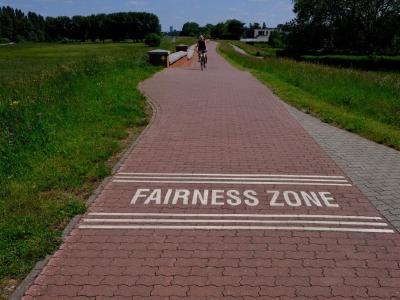 Cyclist on red‑paved path marked “FAIRNESS ZONE” between grassy fields.