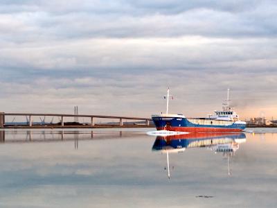 mirrorcalm.jpg A serene waterscape showing a blue and red boat moored near a long wooden pier or bridge, with perfect mirror reflections of both the vessel and cloudy sky in the still water.