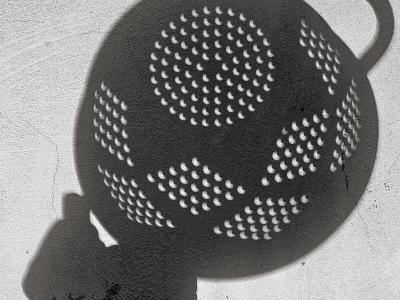 Close-up showing a hand holding a metal colander against a textured off-white wall, with numerous small crescent-shaped eclipse projections visible through the perforations—illustrating the pinhole projection method used for safe solar eclipse viewing.