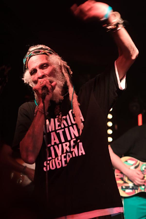 Cedric Myton performing with dramatic lighting, wearing the &ldquo;AMERICA LATINA LIVRE E SOBERANA&rdquo; t-shirt and colorful headband. The image captures him mid-performance with his arm raised in a dynamic blur of motion, microphone in hand.