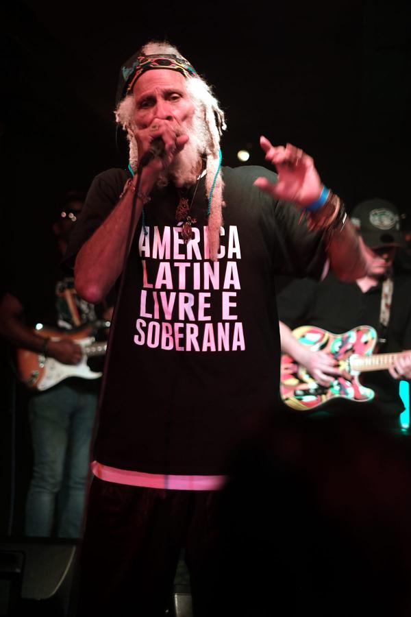 Cedric Myton of The Congos performing on stage wearing a black t-shirt with pink text reading &ldquo;AMERICA LATINA LIVRE E SOBERANA&rdquo; (Free and Sovereign Latin America). He holds a microphone and wears a colorful headband with white dreadlocks visible, his arm raised energetically.