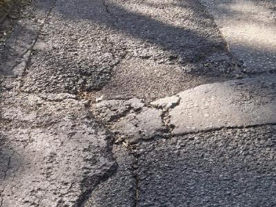 Large pothole with surrounding radial cracking on a sunlit section of Labour in Vain Road, casting long shadows across the damaged pavement.